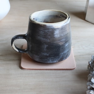 May include: A handmade ceramic mug with a dark, textured exterior and a white rim, resting on a square, light pink leather coaster. The mug has a curved handle and sits on a wooden surface.