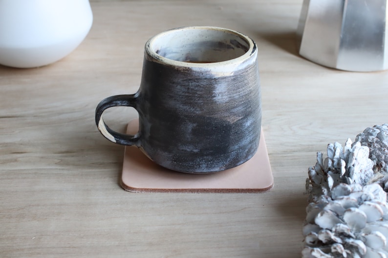 May include: A handmade ceramic mug with a dark, textured exterior and a white rim, resting on a square, light pink leather coaster. The mug has a curved handle and sits on a wooden surface.