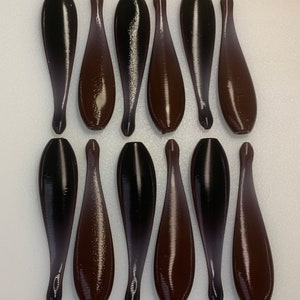 May include: Twelve dark brown, teardrop-shaped fishing lures arranged in two rows of six. Each lure features a smooth, rounded top section that tapers into a thin, elongated tail, with a distinct horizontal ribbed texture across the entire surface.