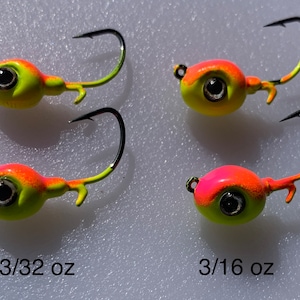 May include: Four fishing jig heads with black hooks. The jig heads are painted with a yellow and orange color scheme. The jig heads are labeled 3/32 oz and 3/16 oz.