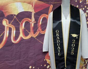 May include: Black graduation sash with gold trim and the words "GRADUATE" and "2025" printed on it. The sash is worn over a white gown.