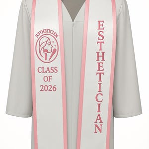 May include: A white graduation gown with a matching sash. The sash has pink trim and the word "ESTHETICIAN" in pink. The sash also features a pink logo and the words "CLASS OF 2026".