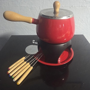 May include: A red fondue pot with a silver lid and wooden handle. The pot sits on a black stand with a red base. Six fondue forks with wooden handles and colored tips are included.