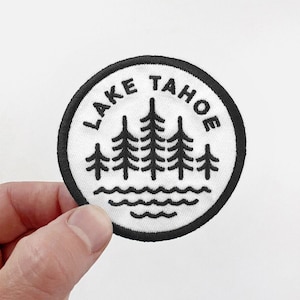 May include: White embroidered patch with black text that reads "LAKE TAHOE" and a design of five pine trees and wavy lines representing water.