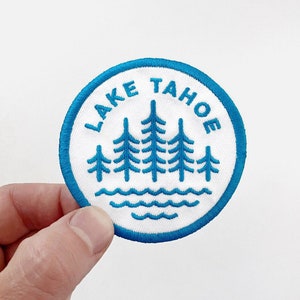 May include: Embroidered patch featuring a blue and white design. The patch has a circular shape with the words "LAKE TAHOE" in blue lettering around the edge. Inside, there are stylized blue trees and water. The patch is held by a hand.