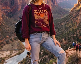 May include: A long-sleeve maroon sweatshirt featuring a graphic print of Zion National Park with a sun and canyon landscape. The sweatshirt is paired with light blue distressed denim jeans, a black backpack, and brown hiking boots.