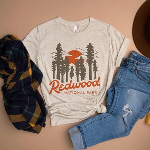 May include: A beige t-shirt with a graphic of a sunset and redwood trees. The text "Redwood National Park" is printed below the graphic.