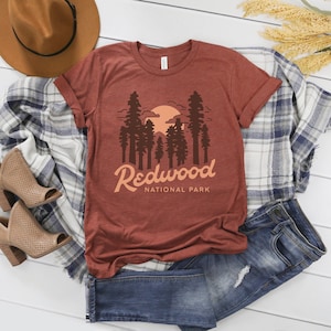 Pode incluir: Uma camiseta cor de ferrugem com um gráfico de um pôr do sol atrás de uma fileira de sequoias. O texto "Redwood National Park" é impresso abaixo do gráfico.
