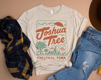 May include: A beige t-shirt with a vintage-style graphic of the Joshua Tree National Park logo. The logo features a red sun, green trees, and the text "Joshua Tree National Park".