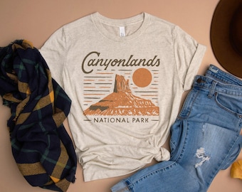 May include: A beige t-shirt with a graphic design of a red rock formation and the text "Canyonlands National Park". The shirt is paired with blue jeans and a brown hat.