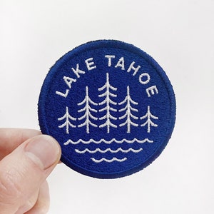 Puede incluir: Un parche circular bordado azul con costuras blancas. El parche presenta un diseño de pinos y olas con el texto "LAKE TAHOE" alrededor del borde.