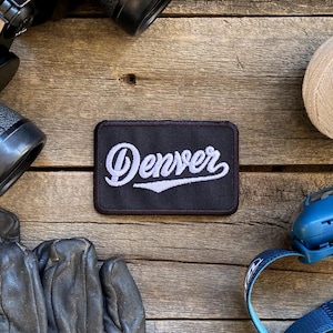 May include: A black and white embroidered patch with the word "Denver" in a white cursive font on a black background. The patch is on a wooden surface with other items, including a pair of binoculars, a spool of twine, and a headlamp.