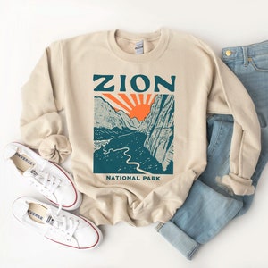 Op de afbeelding: Een beige sweatshirt met een grafische print van een berglandschap en de tekst "Zion National Park".