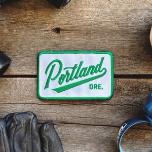 May include: A white patch with green embroidered text that reads "Portland, ORE." The patch is on a wooden surface with other objects.