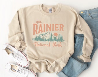 May include: A beige sweatshirt with a graphic print of a mountain range and the text "Mt. Rainier National Park" in a vintage style.