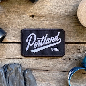 May include: A black and white embroidered patch with the text "Portland, ORE." in a white cursive font on a black background.