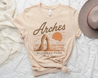 May include: A beige t-shirt with a graphic design of Arches National Park in Moab, Utah. The design features a sunset, a mountain arch, and the text "Arches National Park, Moab, Utah".