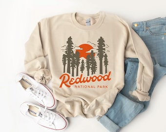May include: A beige sweatshirt with a graphic design of a sunset over a redwood forest. The text "Redwood National Park" is printed below the graphic.
