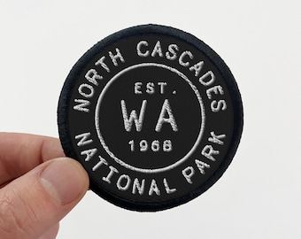May include: Black embroidered patch with white text that reads "North Cascades National Park WA Est. 1968"