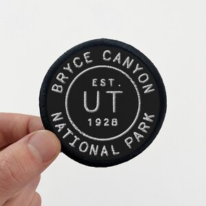 May include: A round black patch with white embroidered text. The text reads "BRYCE CANYON NATIONAL PARK" around the edge, with "UT EST. 1928" in the center. The patch has a black border.