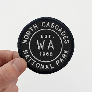May include: Black embroidered patch with white text that reads "North Cascades National Park WA Est. 1968"