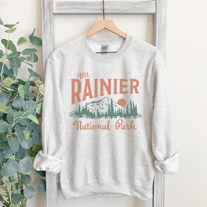 May include: A gray sweatshirt with a vintage-style graphic of Mt. Rainier National Park. The graphic features a mountain range, a setting sun, and trees in shades of green and orange.