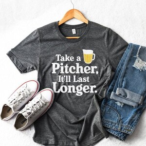 Puede incluir: Una camiseta gris oscuro con un gráfico blanco que dice "Take a Pitcher, It'll Last Longer." El gráfico también incluye una jarra de cerveza amarilla.