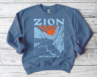 May include: A blue sweatshirt with a graphic of a mountain range and a red sun with the text "ZION NATIONAL PARK" printed on the front.