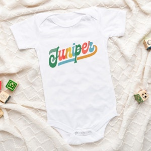 May include: White baby onesie with the name "Juniper" written in colorful retro-style lettering. The onesie is lying on a white knitted blanket with wooden blocks scattered around it.