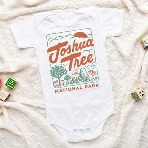 May include: White baby onesie with a green, orange, and brown graphic of Joshua Tree National Park. The text "Joshua Tree National Park" is printed on the onesie.