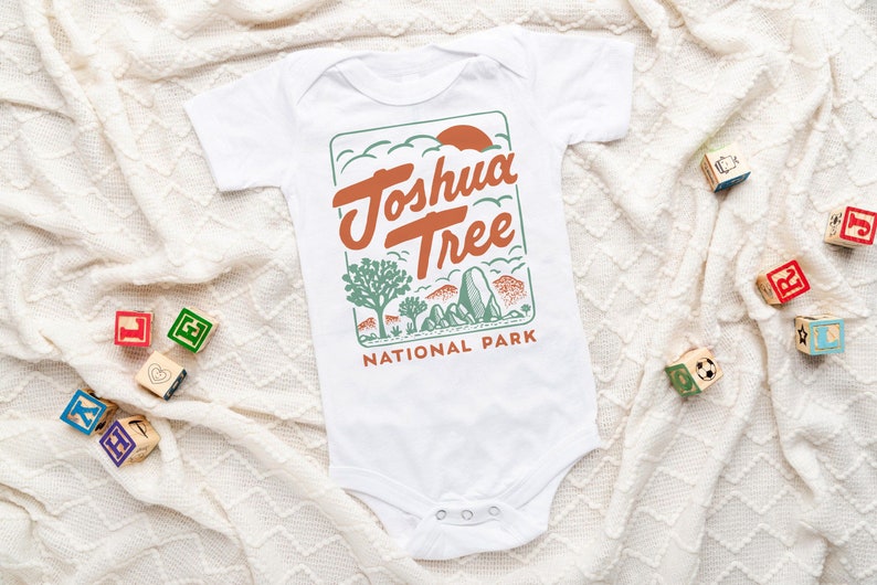 May include: White baby onesie with a green, orange, and brown graphic of Joshua Tree National Park. The text "Joshua Tree National Park" is printed on the onesie.