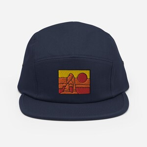 Puede incluir: Una gorra de 5 paneles azul marino con una imagen bordada de una formaci&oacute;n rocosa roja con un sol amarillo en el fondo.