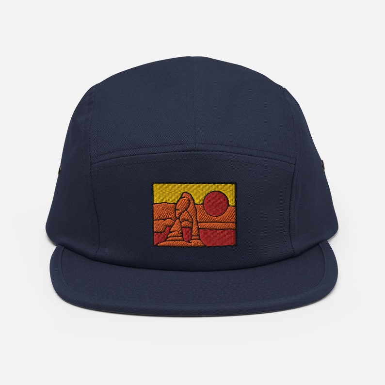 Puede incluir: Una gorra de 5 paneles azul marino con una imagen bordada de una formaci&oacute;n rocosa roja con un sol amarillo en el fondo.