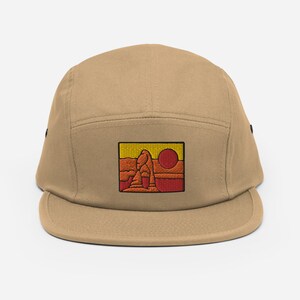 Puede incluir: Una gorra de 5 paneles de color beige con un parche bordado de un paisaje des&eacute;rtico rojo y amarillo con un sol.