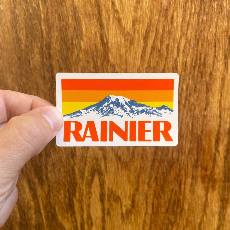May include: A white sticker with a retro-style graphic of Mount Rainier, a mountain with snow-capped peaks, against a sunset background with orange, yellow, and red stripes. The text "RAINIER" is printed in bold, red letters below the mountain.