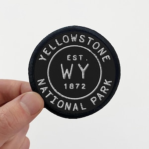May include: A circular embroidered patch with a black background and white border. The text reads YELLOWSTONE NATIONAL PARK in a circular arc, with EST. WY 1872 centered in the middle. The patch features a clean, stitched edge finish.