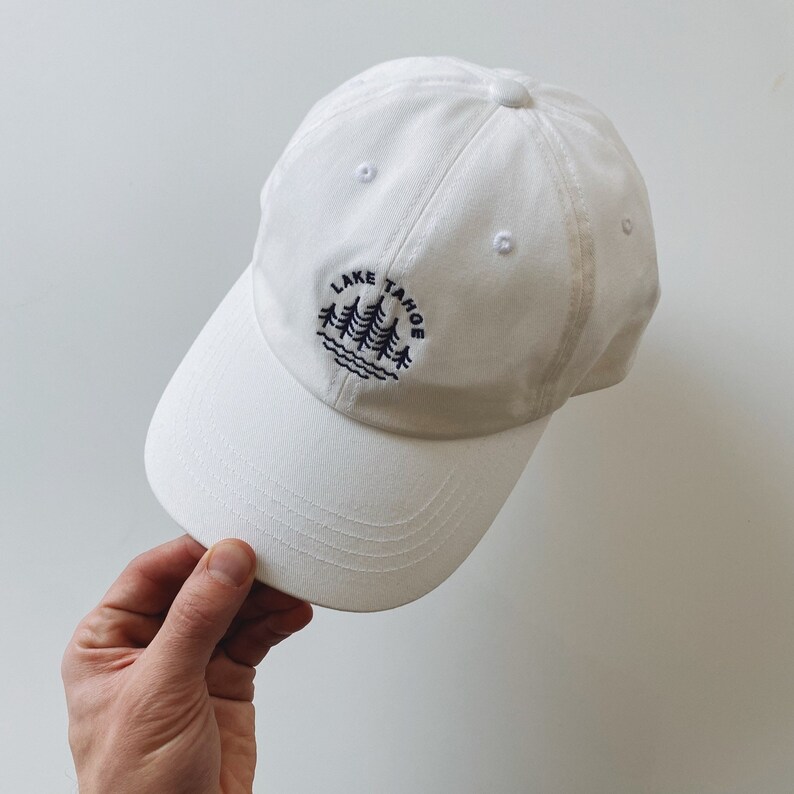 May include: White baseball cap with an embroidered design of pine trees and the text "LAKE TAHOE" in navy blue.