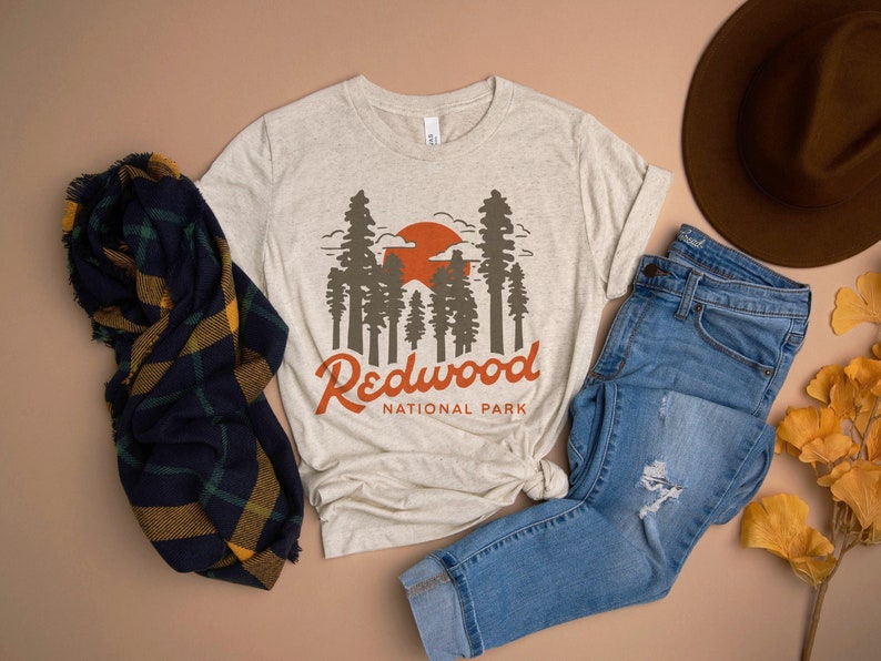 May include: A beige t-shirt with a graphic of a sunset and redwood trees. The text "Redwood National Park" is printed below the graphic.