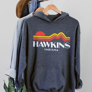 May include: A dark gray hooded sweatshirt with a retro graphic design featuring a sunset over mountains and the text "Hawkins Indiana".