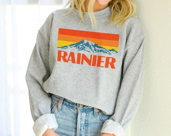 May include: A gray sweatshirt with a retro-style graphic of a mountain range with the word "RAINIER" printed below it. The graphic features a sunset color palette with orange, yellow, and blue hues.