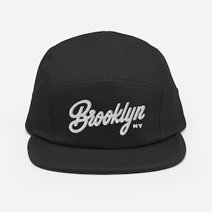 May include: Black 5-panel cap with a white embroidered logo that says "Brooklyn NY" in a cursive font.
