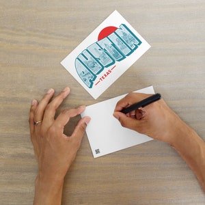 May include: A white postcard with a blue and white illustration of the word "Austin" with a red circle above the "A". The text "Texas" is below the word "Austin". A hand is writing on a blank white postcard with a black pen.