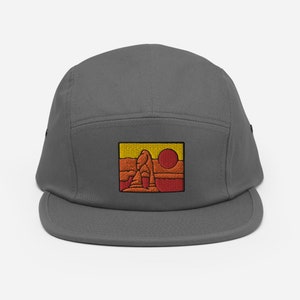 Puede incluir: Una gorra gris de 5 paneles con una imagen bordada de una formaci&oacute;n rocosa roja con un sol amarillo en el fondo.