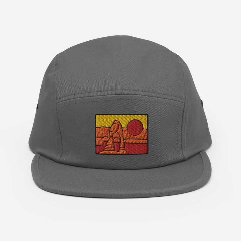 Puede incluir: Una gorra gris de 5 paneles con una imagen bordada de una formaci&oacute;n rocosa roja con un sol amarillo en el fondo.