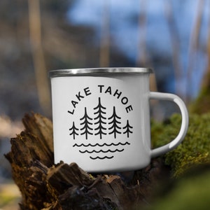 May include: White enamel camping mug with a black graphic of pine trees and the text "LAKE TAHOE" around the trees. The mug is sitting on a tree stump.