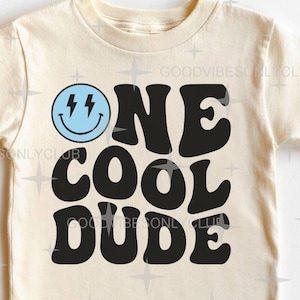 May include: A cream-coloured t-shirt featuring the phrase "ONE COOL DUDE" in bold black lettering. Above "ONE" is a blue smiley face with lightning bolt eyes. This is a casual wear item.