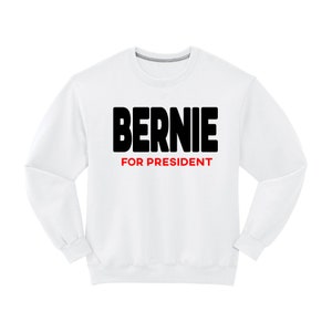 May include: White crew neck sweatshirt featuring the word "BERNIE" in bold black letters above the words "FOR PRESIDENT" in red. The sweatshirt has long sleeves and a ribbed neckline.