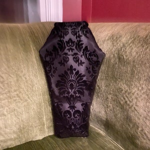 May include: A black velvet coffin-shaped pillow with a damask pattern. The pillow is sitting on a green couch.