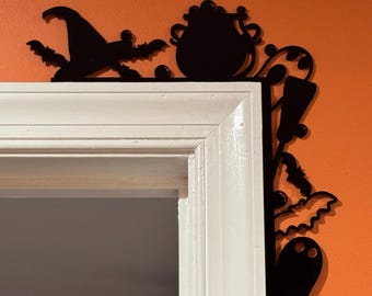 May include: Black Halloween-themed door frame decoration. The silhouette features a witch's hat, cauldron, bats, and a ghost. The decoration is placed on a white door frame against an orange wall.