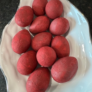 May include: A collection of red decorative eggs with a textured surface, arranged in a white, leaf-shaped ceramic bowl. The eggs are a deep red color, and the bowl has a silver trim.